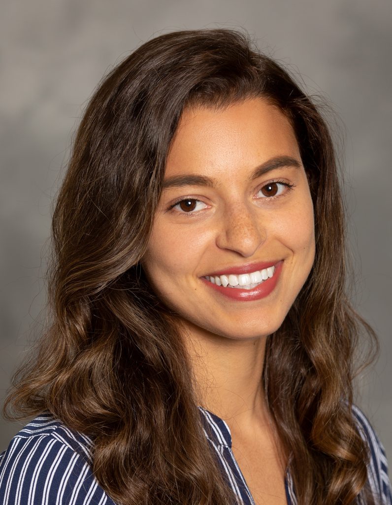 a portrait of Sajadah Connacher looking into the camera and smiling in front of a gray background