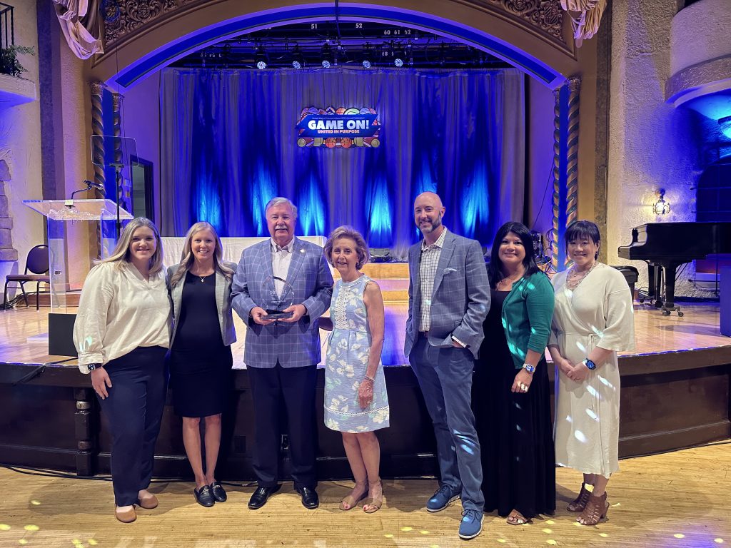 7 people standing in front of a stage. The third person from the left who is president and ceo Jim roe is holding an award.